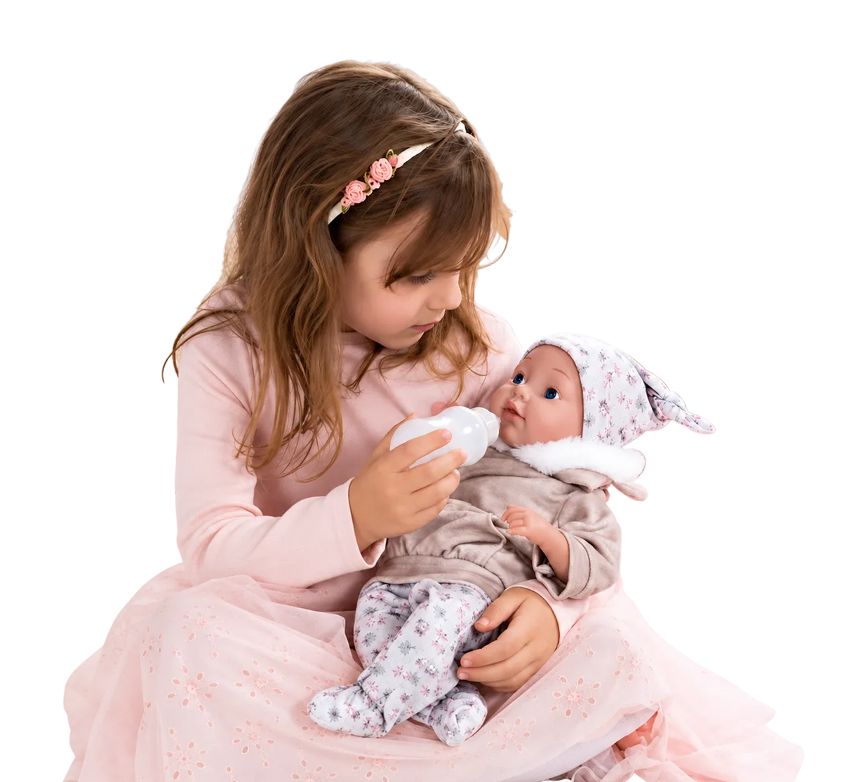 A young girl in a pink dress sits on a bed holding a baby doll dressed in a gray outfit and white floral hat, while offering a bottle. 
